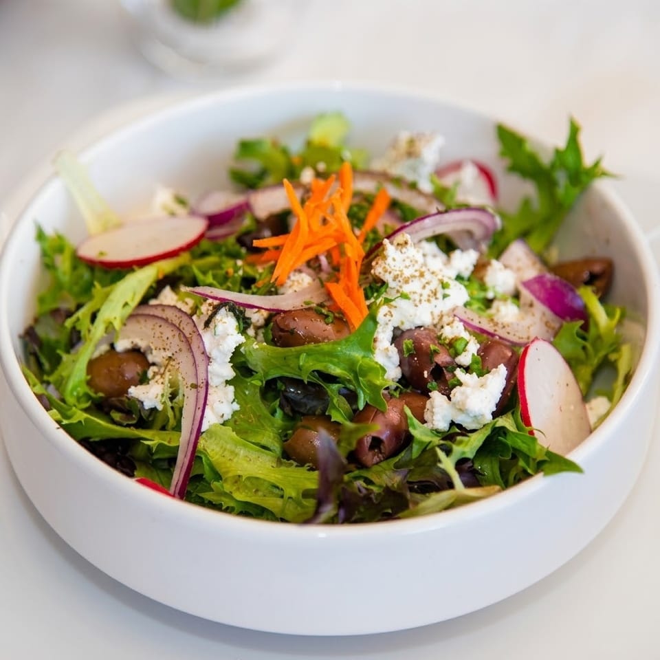 Fresh mixed green salad with olives, feta cheese, sliced radishes, red onion, and shredded carrots in a white bowl.
