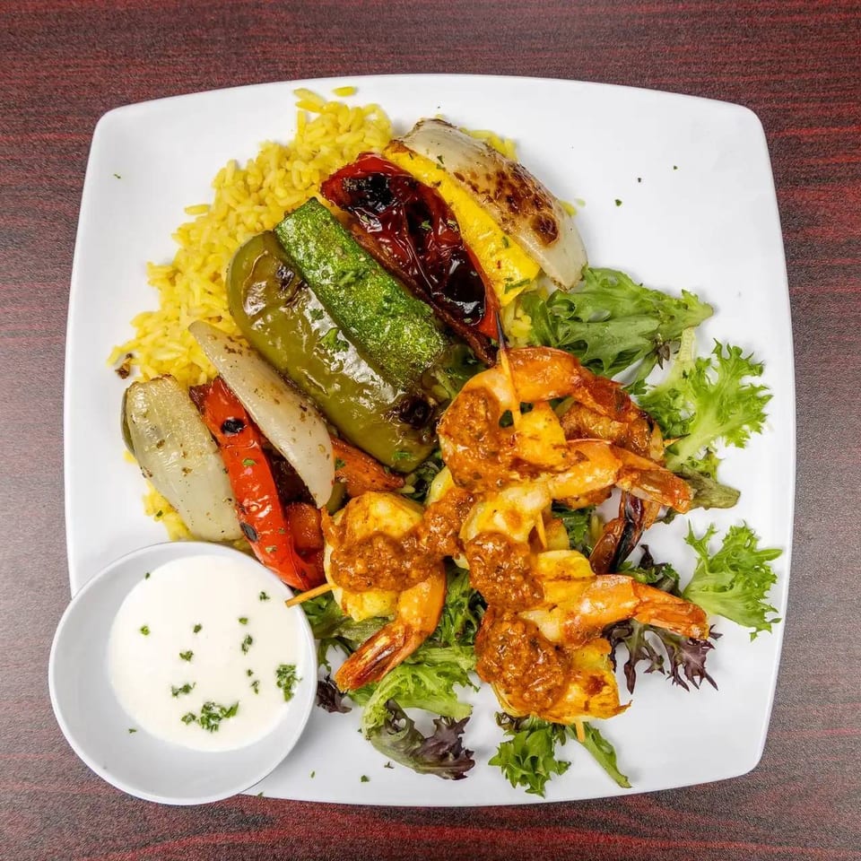 Grilled shrimp with roasted vegetables, yellow rice, mixed greens, and creamy herb sauce on a white plate.