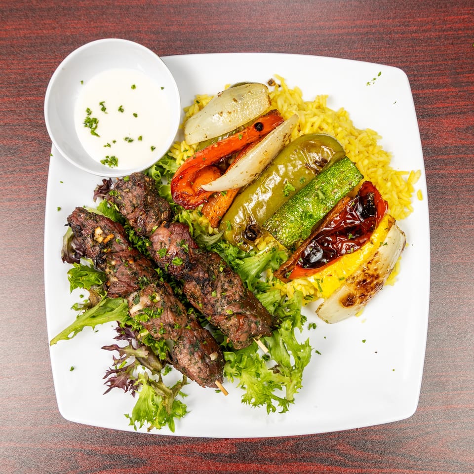 Grilled beef skewers, roasted vegetables, and yellow rice with creamy herb sauce on a white plate.