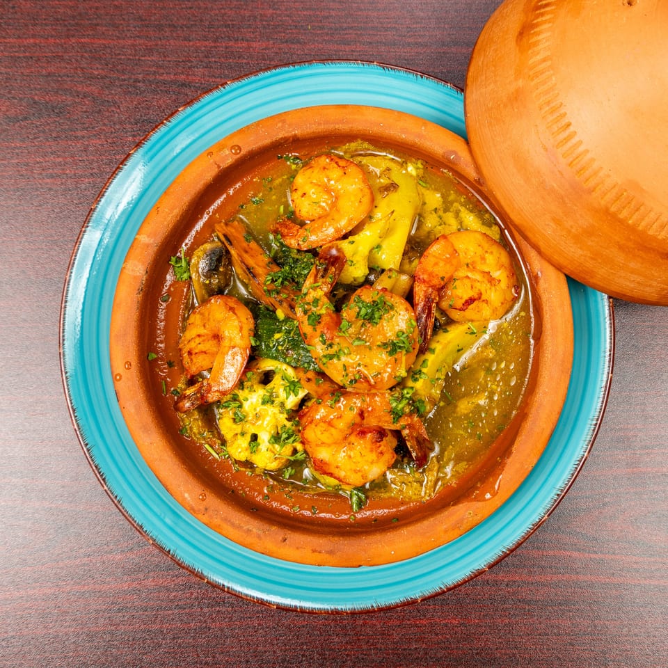 Shrimp and vegetables garnished with herbs in a rustic clay tagine dish on a turquoise plate.